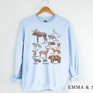 May include: Light blue sweatshirt with a graphic print of various woodland animals, including a moose, wolf, deer, fox, squirrel, raccoon, badger, hedgehog, bear, hare, chipmunk, and fawn. The text "EMMA & ANNA" is printed on the bottom right corner of the sweatshirt.