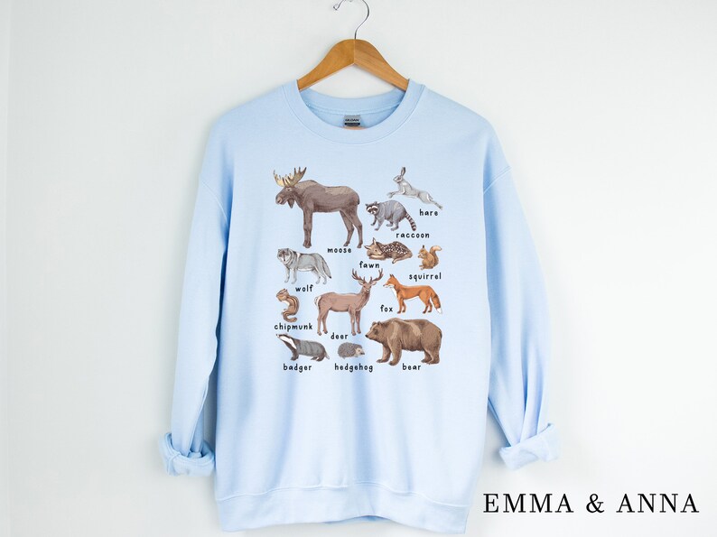 May include: Light blue sweatshirt with a graphic print of various woodland animals, including a moose, wolf, deer, fox, squirrel, raccoon, badger, hedgehog, bear, hare, chipmunk, and fawn. The text "EMMA & ANNA" is printed on the bottom right corner of the sweatshirt.