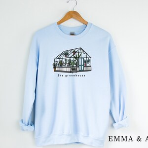May include: Pale blue sweatshirt with a graphic of a greenhouse, complete with plants and the words "the greenhouse". The design is in black and green. The sweatshirt is displayed on a wooden hanger.