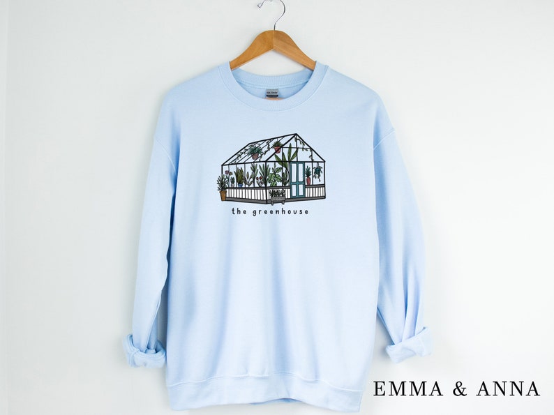 May include: Pale blue sweatshirt with a graphic of a greenhouse, complete with plants and the words "the greenhouse". The design is in black and green. The sweatshirt is displayed on a wooden hanger.