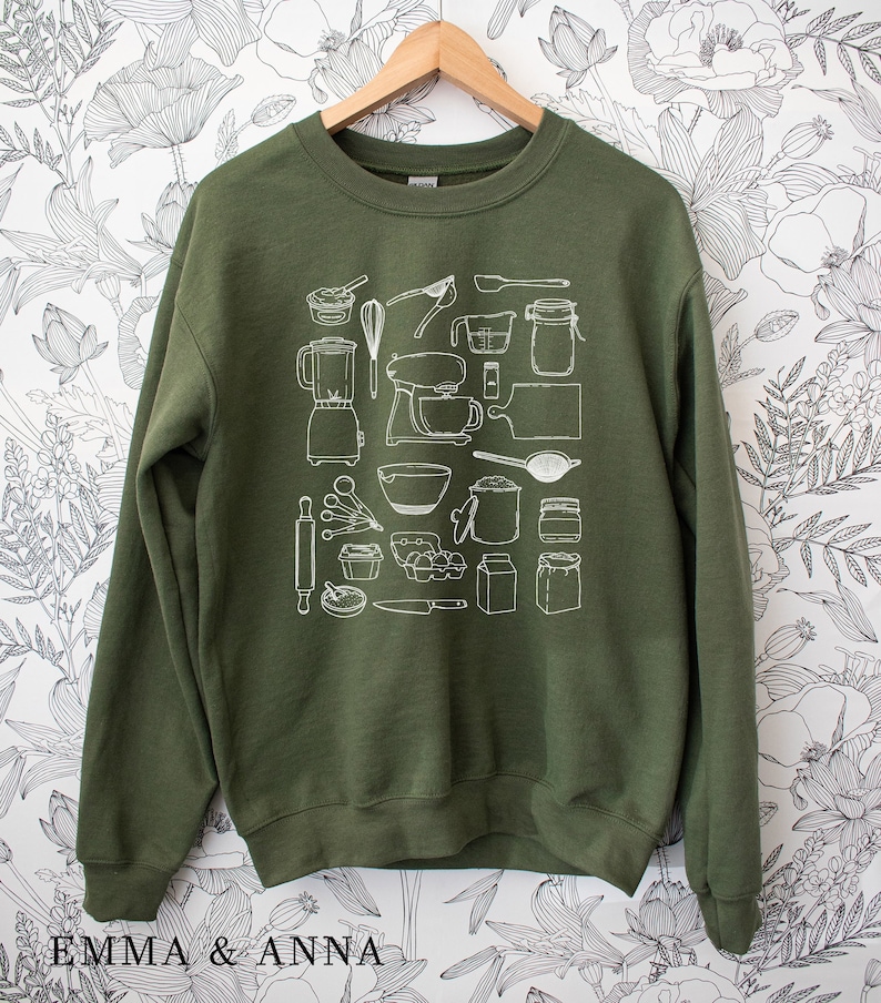 May include: A green sweatshirt with a white graphic print of various kitchen tools and appliances. The graphic includes a blender, mixer, rolling pin, measuring cups, and more.