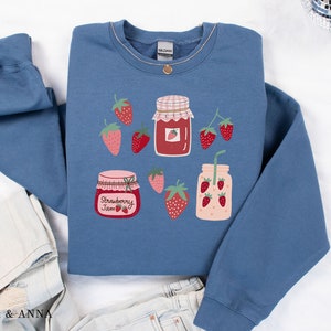 May include: A blue sweatshirt with a strawberry jam jar and a strawberry smoothie design. The jars have white lids and red strawberry designs. The text "Strawberry Jam" is printed on one of the jars.
