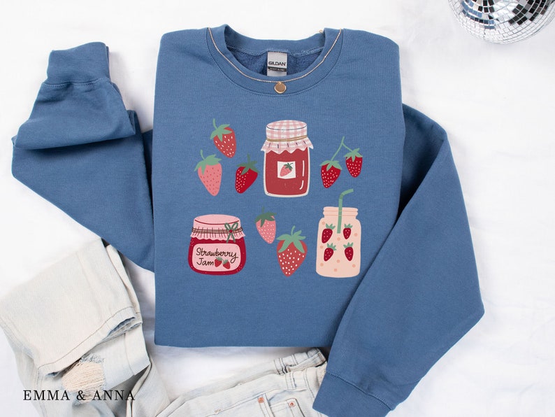 May include: A blue sweatshirt with a strawberry jam jar and a strawberry smoothie design. The jars have white lids and red strawberry designs. The text "Strawberry Jam" is printed on one of the jars.