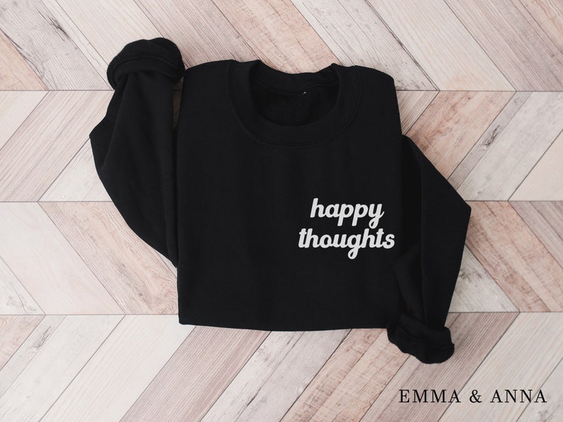 May include: Black sweatshirt with the text "happy thoughts" in white on the front. The sweatshirt is folded and laid flat on a woodgrain surface.