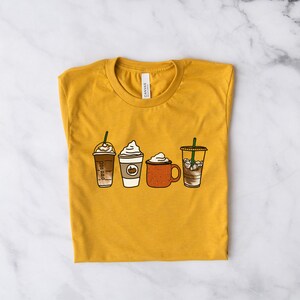 Puede incluir: Una camiseta amarilla mostaza con un gr&aacute;fico de cuatro bebidas de caf&eacute; diferentes. Las bebidas incluyen un caramel macchiato, un latte de especias de calabaza, un chocolate caliente y un caf&eacute; helado.