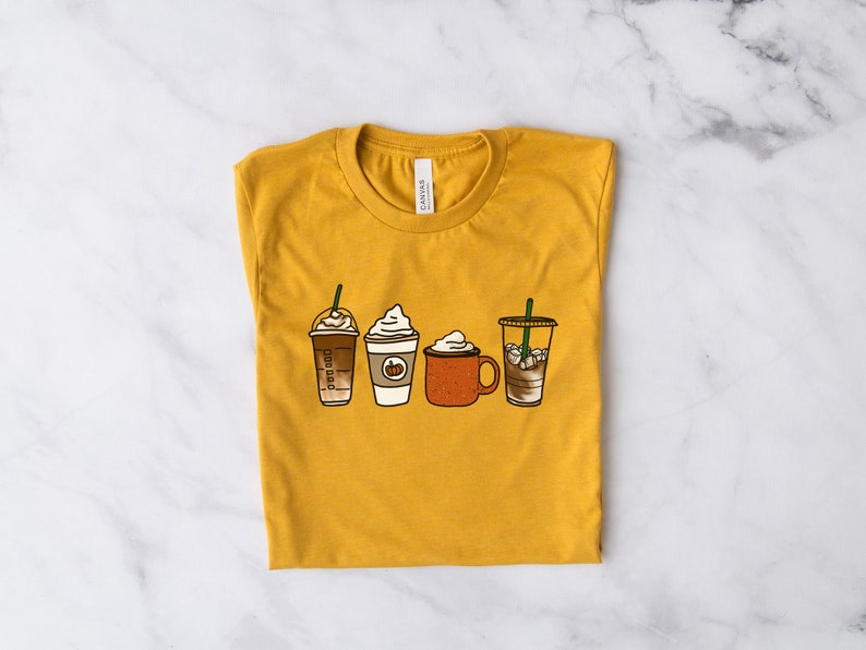 Puede incluir: Una camiseta amarilla mostaza con un gr&aacute;fico de cuatro bebidas de caf&eacute; diferentes. Las bebidas incluyen un caramel macchiato, un latte de especias de calabaza, un chocolate caliente y un caf&eacute; helado.