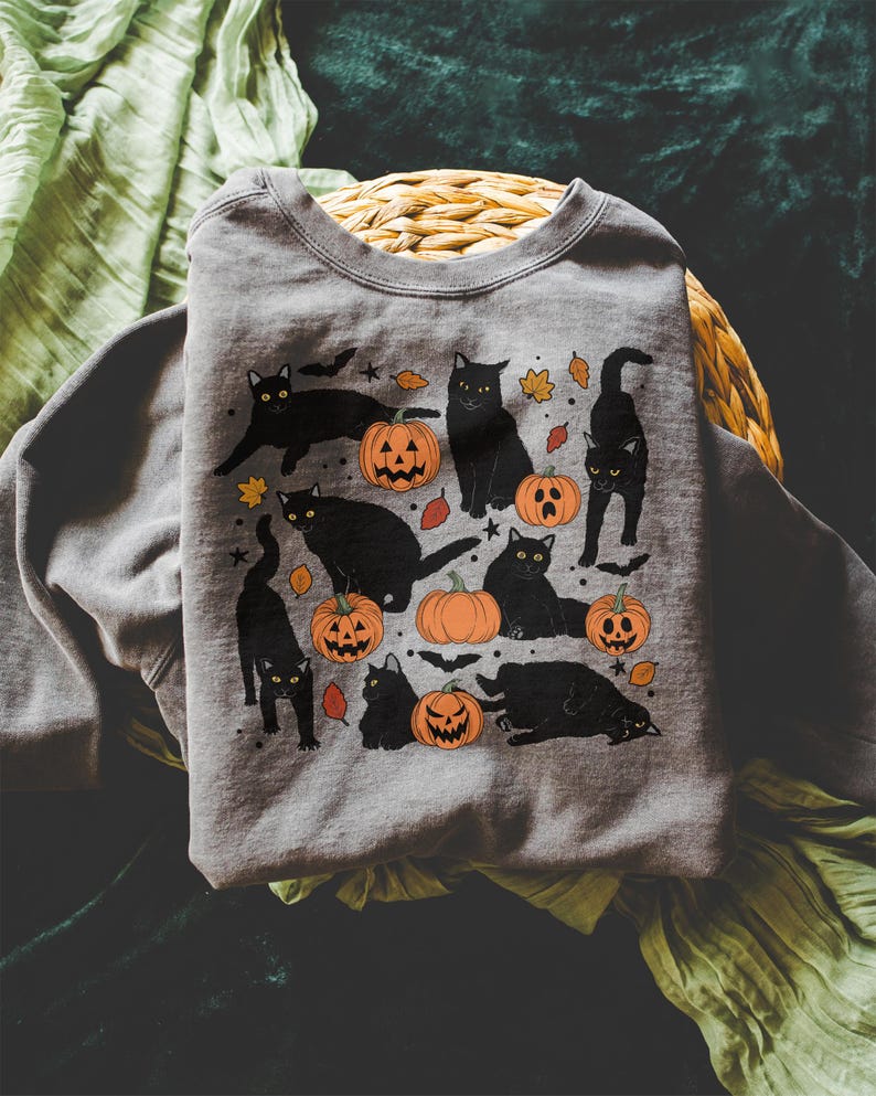 May include: A gray sweatshirt featuring a Halloween-themed design. The graphic includes black cats, orange pumpkins with carved faces, and autumn leaves. The cats are in various poses, creating a playful and festive look.