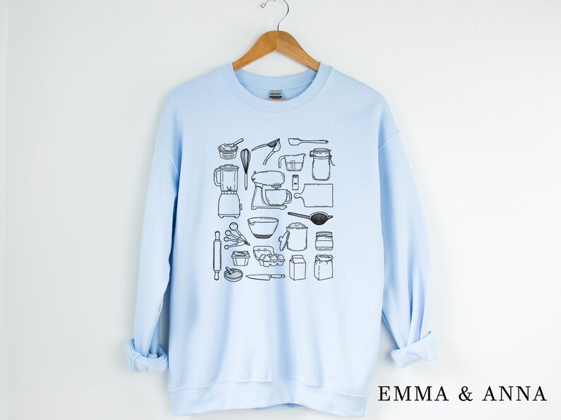 May include: A light blue sweatshirt with a black and white illustration of various baking tools and ingredients. The text "EMMA & ANNA" is printed on the bottom right corner of the sweatshirt.