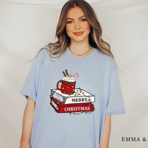 May include: Light blue t-shirt with a graphic of a red mug with a white whipped cream topping, a candy cane, and a stack of books with the text "Merry Christmas" on the front. The graphic is surrounded by red and green Christmas lights.
