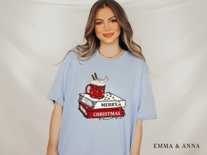 May include: Light blue t-shirt with a graphic of a red mug with a white whipped cream topping, a candy cane, and a stack of books with the text "Merry Christmas" on the front. The graphic is surrounded by red and green Christmas lights.