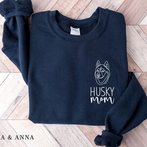 Pode incluir: Uma camisola azul marinho com um contorno branco de um c&atilde;o husky e o texto "HUSKY MOM" na frente.