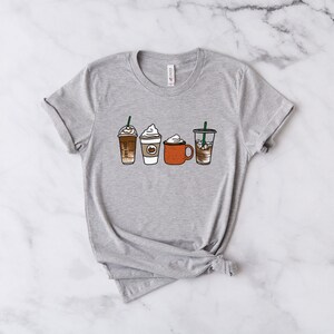 Puede incluir: Una camiseta gris con un gr&aacute;fico de cuatro bebidas de caf&eacute; diferentes, incluyendo un latte, un capuchino, un chocolate caliente y un caf&eacute; helado.