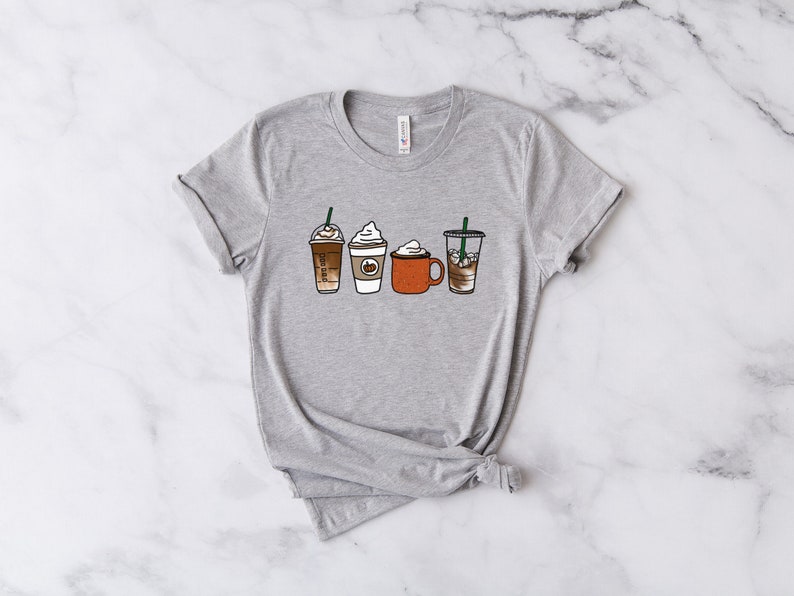 Puede incluir: Una camiseta gris con un gr&aacute;fico de cuatro bebidas de caf&eacute; diferentes, incluyendo un latte, un capuchino, un chocolate caliente y un caf&eacute; helado.