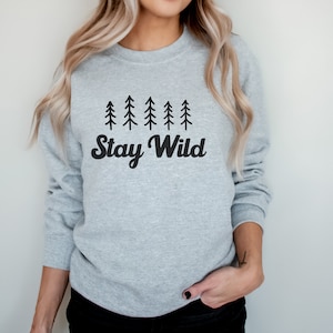 K&ouml;nnte beinhalten: Ein grauer Pullover mit einer schwarzen Grafik von f&uuml;nf stilisierten Kiefern und dem Text "Stay Wild".