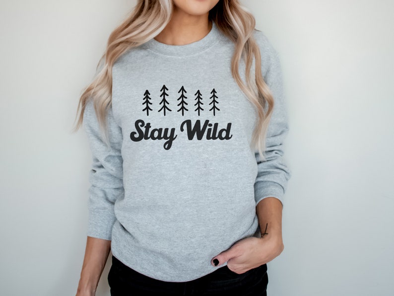 K&ouml;nnte beinhalten: Ein grauer Pullover mit einer schwarzen Grafik von f&uuml;nf stilisierten Kiefern und dem Text "Stay Wild".