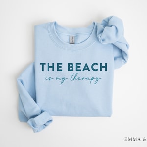 以下が含まれることがあります： 薄い青色のスウェットシャツに、濃い青色の文字で「THE BEACH is my therapy」と書かれています。