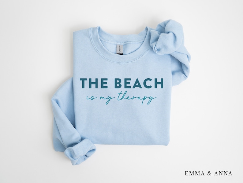 以下が含まれることがあります： 薄い青色のスウェットシャツに、濃い青色の文字で「THE BEACH is my therapy」と書かれています。
