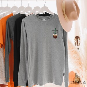 Peut inclure: Un t-shirt &agrave; manches longues gris chin&eacute; avec un motif de boisson au caf&eacute; sur la poitrine gauche. Le t-shirt est suspendu &agrave; un cintre blanc.