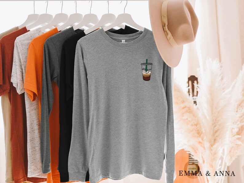 Peut inclure: Un t-shirt &agrave; manches longues gris chin&eacute; avec un motif de boisson au caf&eacute; sur la poitrine gauche. Le t-shirt est suspendu &agrave; un cintre blanc.