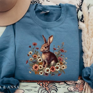 Pu&ograve; includere: Felpa blu con un coniglio marrone e un design floreale. Il coniglio &egrave; circondato da fiori colorati. La felpa ha girocollo e maniche lunghe. Il testo "EMMA & ANNA" &egrave; stampato in basso a sinistra. Un capo comodo e casual.
