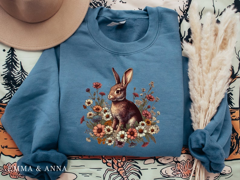 Pu&ograve; includere: Felpa blu con un coniglio marrone e un design floreale. Il coniglio &egrave; circondato da fiori colorati. La felpa ha girocollo e maniche lunghe. Il testo "EMMA & ANNA" &egrave; stampato in basso a sinistra. Un capo comodo e casual.