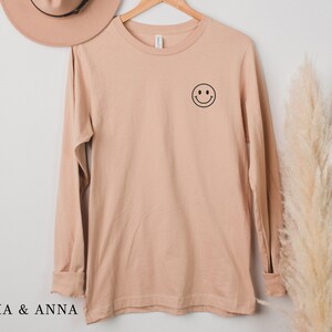 Puede incluir: Una camiseta de manga larga de color beige claro con un peque&ntilde;o smiley negro en la parte superior izquierda del pecho. La camiseta est&aacute; colgada de una percha de madera. El texto "EMMA & ANNA" est&aacute; en la parte inferior de la imagen.