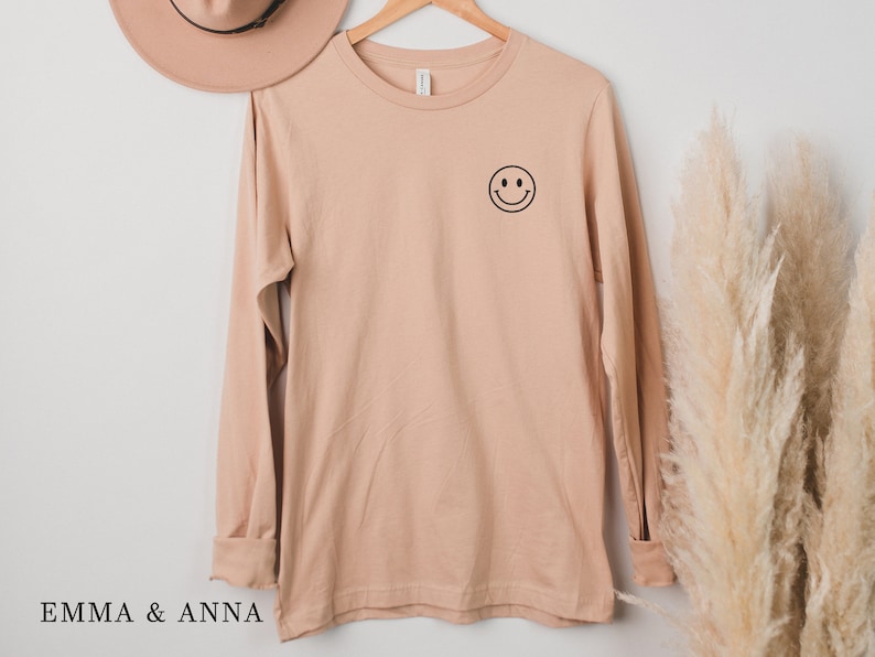 Puede incluir: Una camiseta de manga larga de color beige claro con un peque&ntilde;o smiley negro en la parte superior izquierda del pecho. La camiseta est&aacute; colgada de una percha de madera. El texto "EMMA & ANNA" est&aacute; en la parte inferior de la imagen.