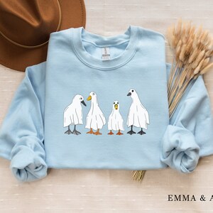 May include: A light blue sweatshirt with a graphic of four white ghost ducks. The ducks are lined up in a row and have black eyes and orange beaks.