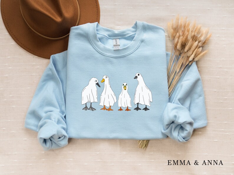 May include: A light blue sweatshirt with a graphic of four white ghost ducks. The ducks are lined up in a row and have black eyes and orange beaks.