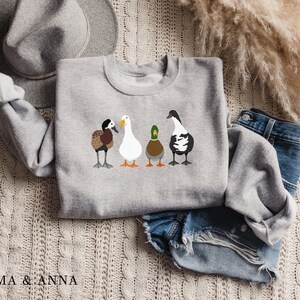 Peut inclure: Un sweat-shirt gris avec un motif de quatre canards align&eacute;s. Les canards sont de couleurs diff&eacute;rentes et ont des marques diff&eacute;rentes. Le sweat-shirt est pos&eacute; sur une surface en tricot blanc avec un chapeau marron et un jean bleu.