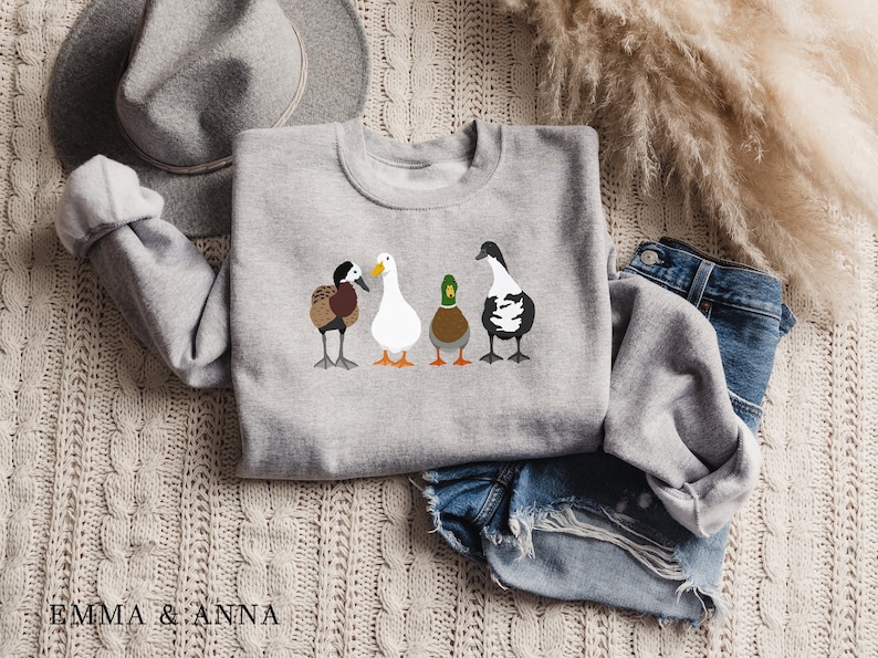 Peut inclure: Un sweat-shirt gris avec un motif de quatre canards align&eacute;s. Les canards sont de couleurs diff&eacute;rentes et ont des marques diff&eacute;rentes. Le sweat-shirt est pos&eacute; sur une surface en tricot blanc avec un chapeau marron et un jean bleu.