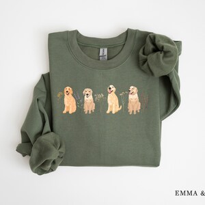May include: A green sweatshirt with a crew neck and a print of four golden retriever dogs in a row. The dogs are standing in a field of flowers.