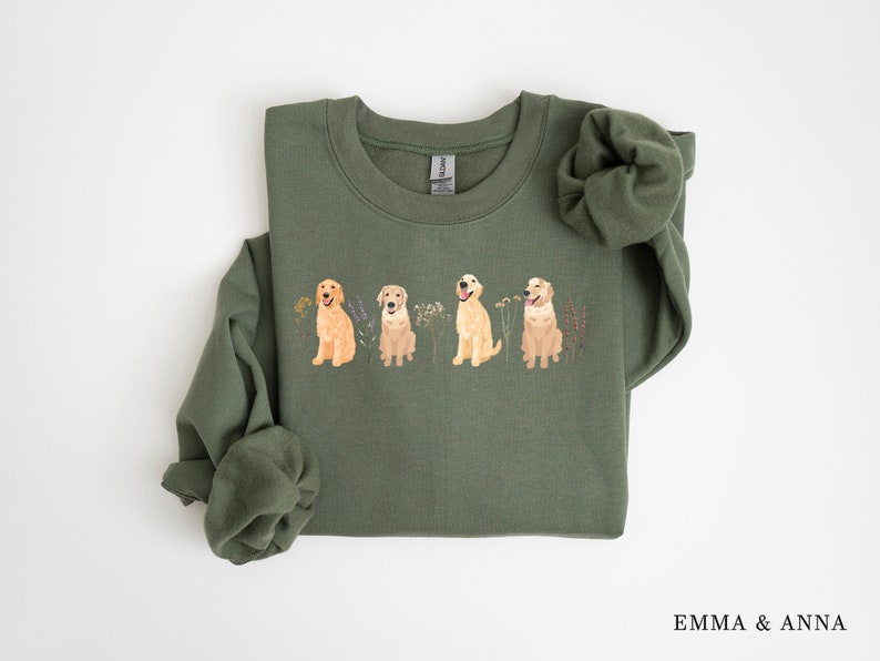 May include: A green sweatshirt with a crew neck and a print of four golden retriever dogs in a row. The dogs are standing in a field of flowers.