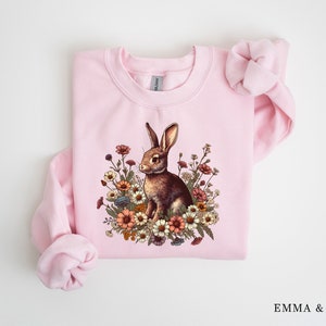 Pu&ograve; includere: Una felpa rosa chiaro con un coniglio marrone circondato da fiori colorati. Il coniglio &egrave; al centro, circondato da fiori. La felpa ha maniche lunghe e girocollo.