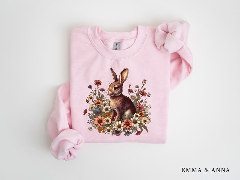 Pu&ograve; includere: Una felpa rosa chiaro con un coniglio marrone circondato da fiori colorati. Il coniglio &egrave; al centro, circondato da fiori. La felpa ha maniche lunghe e girocollo.