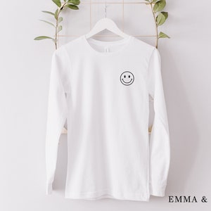 Puede incluir: Una camiseta blanca de manga larga con un peque&ntilde;o gr&aacute;fico de carita sonriente negra en la parte superior izquierda del pecho. La camiseta est&aacute; colgada en una percha blanca sobre un fondo blanco. El texto "EMMA & ANNA" est&aacute; en la parte inferior derecha.