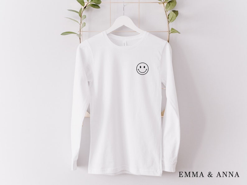 Puede incluir: Una camiseta blanca de manga larga con un peque&ntilde;o gr&aacute;fico de carita sonriente negra en la parte superior izquierda del pecho. La camiseta est&aacute; colgada en una percha blanca sobre un fondo blanco. El texto "EMMA & ANNA" est&aacute; en la parte inferior derecha.