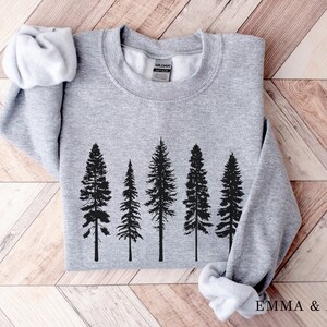 Puede incluir: Una sudadera gris con una silueta negra de cinco pinos impresa en la parte delantera. La sudadera est&aacute; doblada y colocada sobre una superficie de madera.