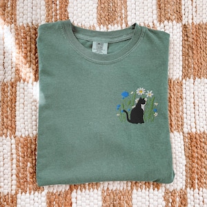 May include: A sage green t-shirt with an embroidered design of a black cat sitting among white and blue flowers. The shirt is folded and lies on a woven, patterned surface.