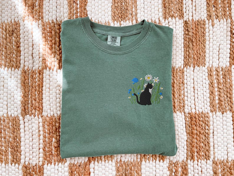 May include: A sage green t-shirt with an embroidered design of a black cat sitting among white and blue flowers. The shirt is folded and lies on a woven, patterned surface.