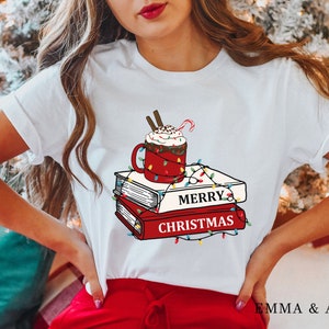 May include: White t-shirt with a graphic of a red mug with whipped cream and a candy cane on top of a stack of books. The books have the text "Merry Christmas" on them and are decorated with colorful lights.