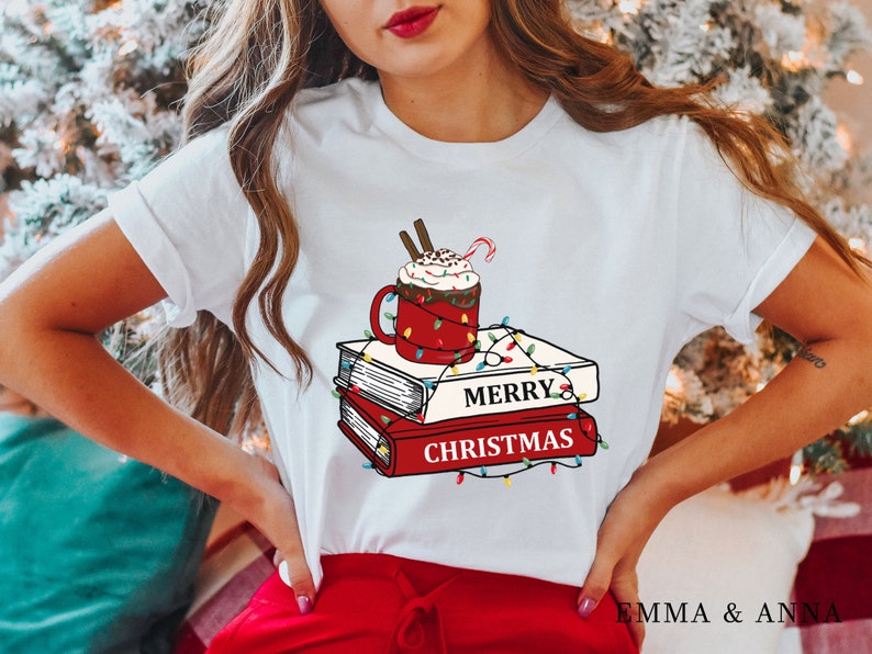 May include: White t-shirt with a graphic of a red mug with whipped cream and a candy cane on top of a stack of books. The books have the text "Merry Christmas" on them and are decorated with colorful lights.