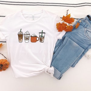 Puede incluir: Camiseta blanca con un gr&aacute;fico de cuatro bebidas de caf&eacute; diferentes, incluyendo un latte, un cappuccino, un latte de especias de calabaza y un caf&eacute; helado. La camiseta se combina con jeans azules y hojas de oto&ntilde;o.