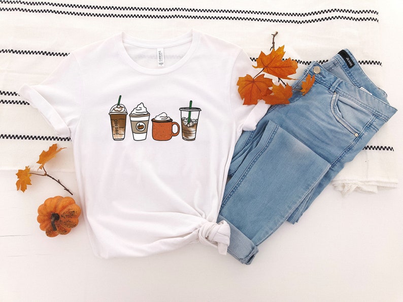 Puede incluir: Camiseta blanca con un gr&aacute;fico de cuatro bebidas de caf&eacute; diferentes, incluyendo un latte, un cappuccino, un latte de especias de calabaza y un caf&eacute; helado. La camiseta se combina con jeans azules y hojas de oto&ntilde;o.