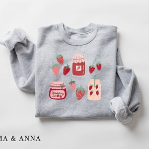 May include: Gray sweatshirt with a strawberry jam jar, a glass of strawberry juice, and strawberries printed on it. The text "Strawberry Jam" is printed on the jar.