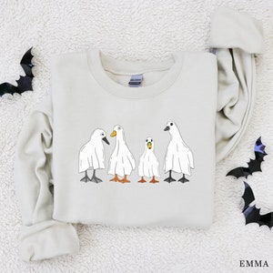 May include: A light beige sweatshirt with a graphic of four white ghost ducks. The ducks are in a line, with the largest duck in the back and the smallest duck in the front. The sweatshirt has a crew neck and long sleeves.