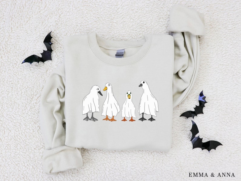 May include: A light beige sweatshirt with a graphic of four white ghost ducks. The ducks are in a line, with the largest duck in the back and the smallest duck in the front. The sweatshirt has a crew neck and long sleeves.