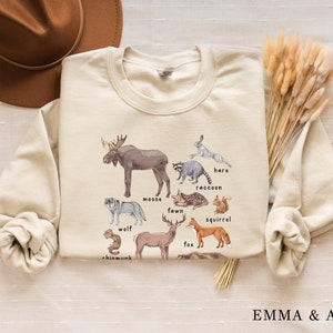 May include: A beige sweatshirt with a graphic print of various forest animals, including a moose, wolf, fawn, fox, squirrel, raccoon, hare, and chipmunk. The animals are illustrated in a watercolor style and labeled with their names.