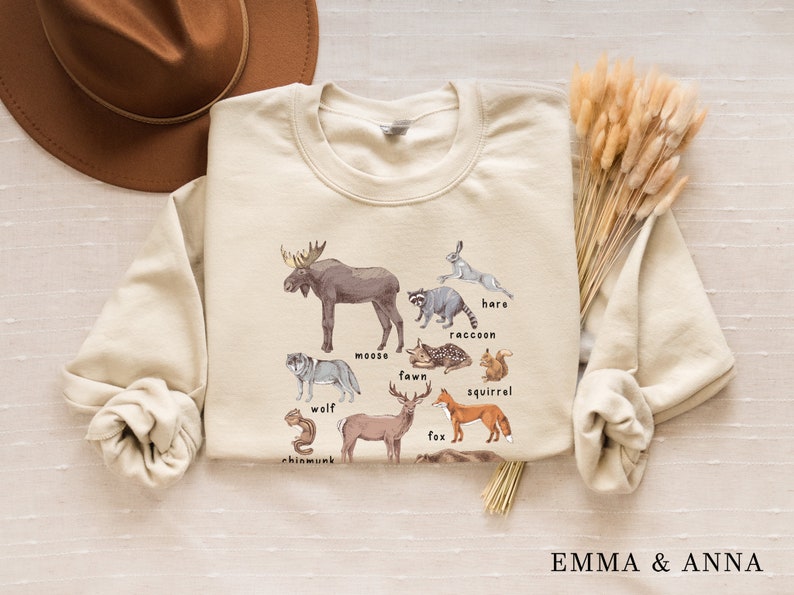 May include: A beige sweatshirt with a graphic print of various forest animals, including a moose, wolf, fawn, fox, squirrel, raccoon, hare, and chipmunk. The animals are illustrated in a watercolor style and labeled with their names.