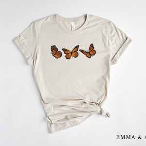 Może przedstawiać: Jasny beżowy t-shirt z trzema ilustracjami pomarańczowo-czarnych motyli monarchy. Motyle ułożone są poziomo na piersi. Tekst "EMMA & ANNA" jest wydrukowany na dole po prawej stronie.